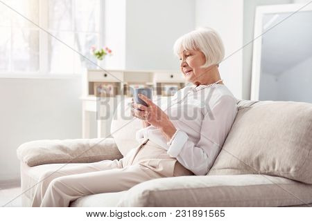 Cozy Morning. Charming Elderly Woman Sitting On The Comfortable Couch, Holding A Cup Of Coffee And L