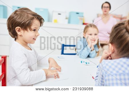 Little elementary school learner listening to one of groupmates at lesson of English language