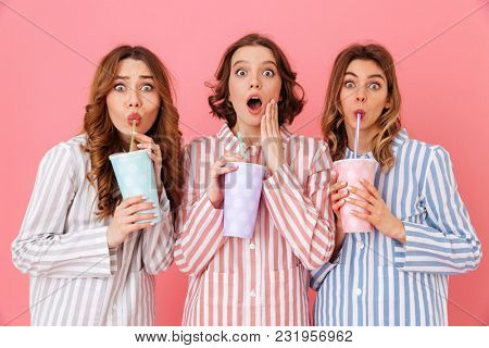 Three brunette girls 20s wearing leisure clothing drinking cold beverages from paper cups and expressing surprise during happy sleepover isolated over pink background