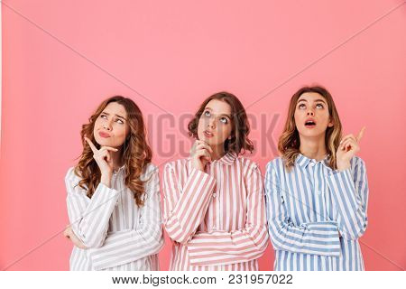 Portrait of three cute young girls 20s wearing colorful striped pyjamas posing on camera with pensive or brooding look during sleepover isolated over pink background