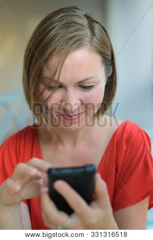 Young Adult Woman In Red Dress Sitting At Cafe Near Window, Looking Down And Internet Surfing In Sma