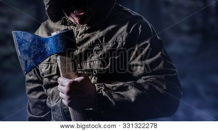 Photo of a scary angry killer male holding an axe in dark foggy woods background.