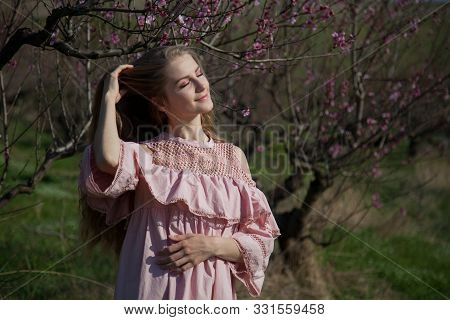 Happy Blonde Woman In Pink Dress Walks Through The Flowering Garden In Spring