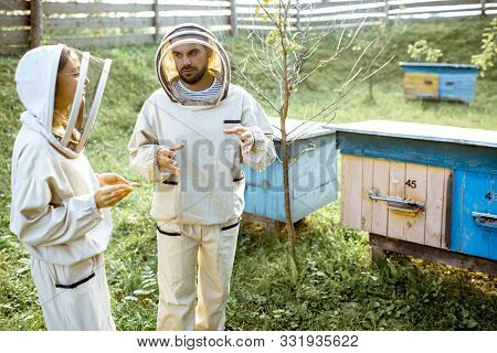 Two Young Beekepers In Protective Uniform Working On A Small Apiary Farm With Wooden Beehives On The