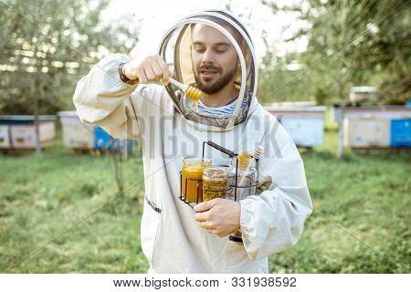 Handsome Beekeper In Protective Uniform Standing With Honey In The Jar, Tasting Fresh Product On The