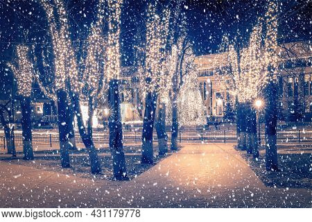 Winter Park At Night With Christmas Decorations, Lights, Pavement Covered Withsnow And Trees.