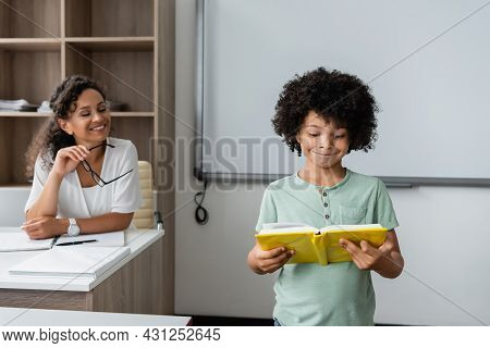 Smiling African American Teacher Looking At Schoolboy Reading Book In Classroom