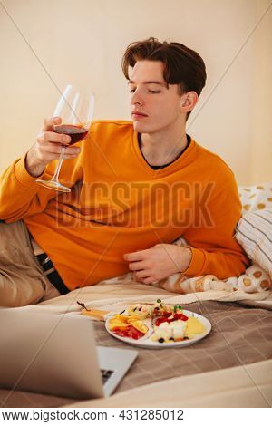 Relaxed Young Guy With Glass Of Wine In His Hands Laying On Comfortable Bed With Plate Of Food, Watc
