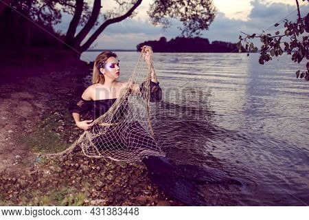 Mermaid Posing With Net Near Sea On Rocks While Wearing Artistic Makeup And Facial Strassses And Bla