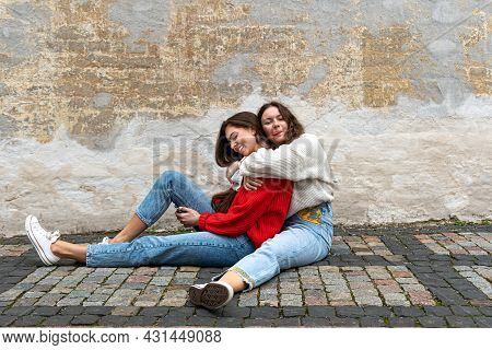 Two Best Friends Hugged Sitting On A Paved Sidewalk By An Old Wall