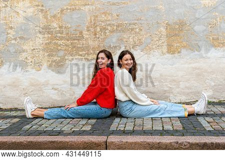 Two Young Women Sitting Back To Back On The Pavement By An Old Wall