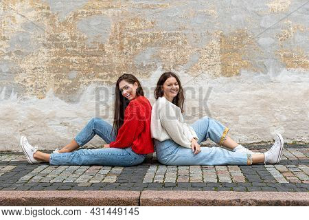 Two Young Women Sitting Back To Back On The Pavement By An Old Wall