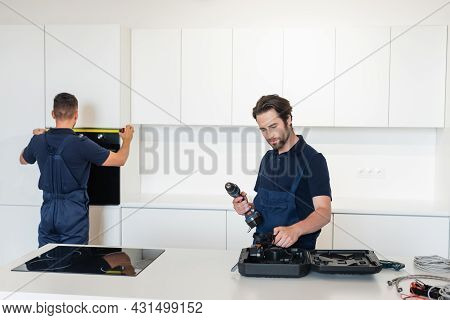 Workman Holding Electric Drill Near Colleague Measuring Inbuilt Oven In Kitchen