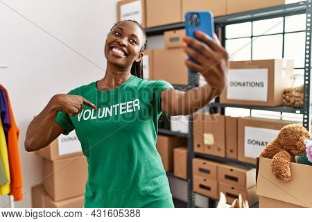 Young african american woman pointing with finger to volunteer tshirt make selfie by the smartphone at charity center.