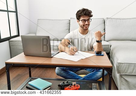 Hispanic man doing papers at home smiling with happy face looking and pointing to the side with thumb up. 