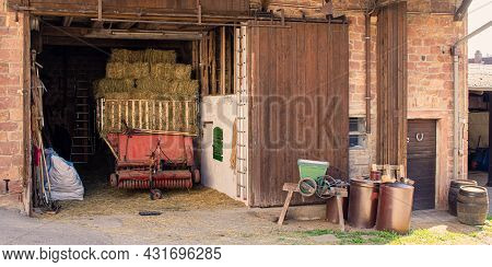Agricultural Objects In Front Of A Barn Of An Old Farmhouse With Grain Crasher, Silage Wagon And Bar