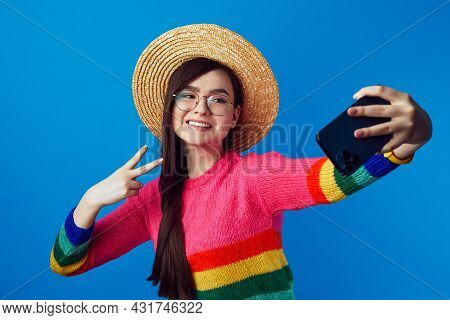 Attractive Female Wearing Rainbow Sweater, Eyeglasses And Hat, Taking Selfie With Smartphone And Sho