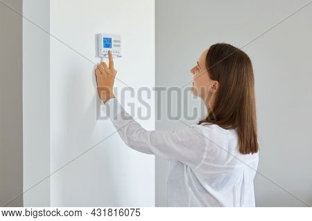 Indoor Shot Of Young Adult Woman Regulating Heating Temperature With A Modern Wireless Thermostat In