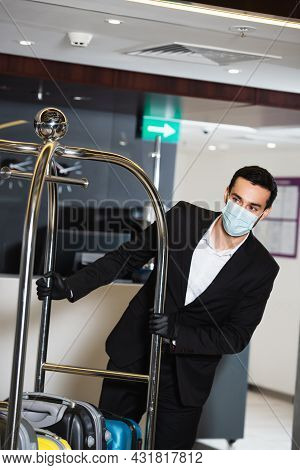 Bellboy In Medical Mask Pushing Luggage Cart In Hotel Lobby