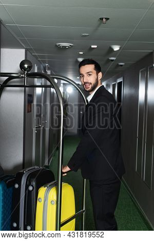 Cheerful Bellboy Near Metallic Bell Cart With Luggage In Hall Of Hotel
