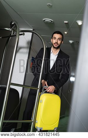 Smiling Bellboy Carrying Luggage Near Metallic Bell Cart In Hall Of Hotel