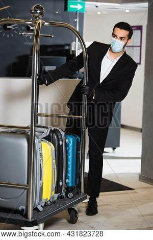 Bellboy In Medical Mask Pushing Bell Cart With Luggage In Hotel Lobby
