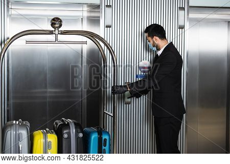 Bellboy In Medical Mask Standing With Bell Cart Near Elevator In Hotel