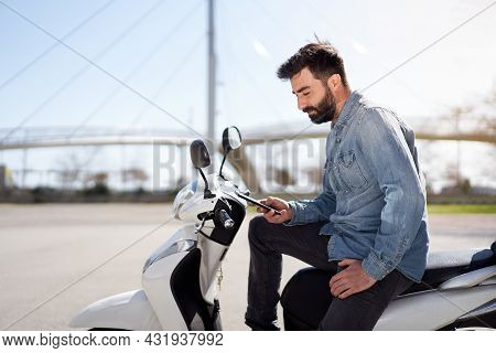 Young Spanish Man Using Cell Phone Or Smartphone Sitting On His Motor Scooter Outdoors In The City. 