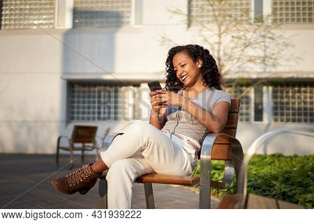Young Beautiful Woman Typing On Cell Phone Sitting On Bench- Smiling Mixed Race Woman Using Smartpho