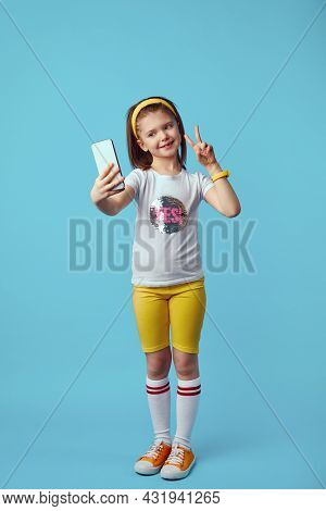 Adorable Kid Girl Takes Selfie Portrait On Mobile Phone, Shows Peace Gesture, Wears White T Shirt, Y