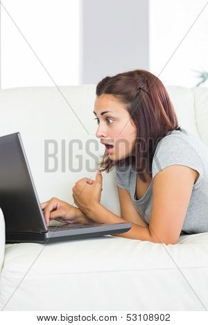 Brunette young woman using her notebook lying taken aback in front of it at home in the living room