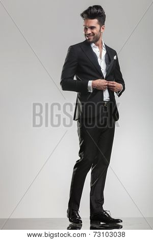 Full body image of an elegant young man closing his tuxedo, smiling while looking away from the camera.