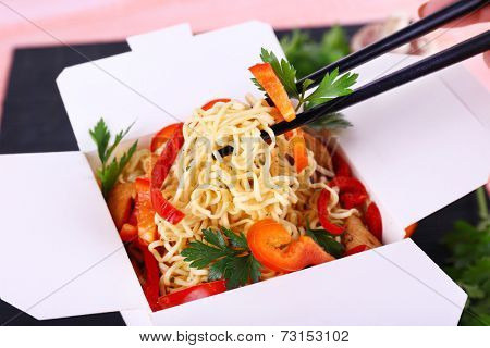Chinese noodles and sticks in takeaway box on black mat on pink background