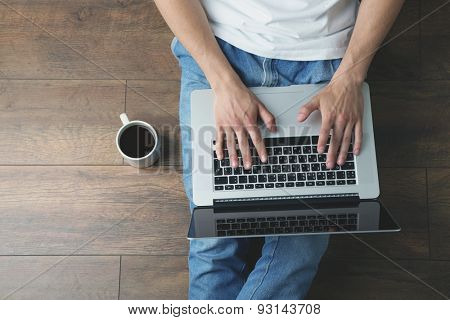 Young man sitting on floor with laptop and cup of coffee in room