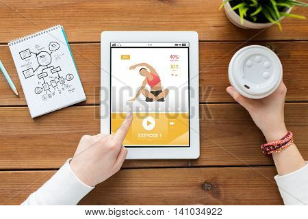 sport, education, technology and people concept - close up of woman with fitness application on tablet pc computer screen, notebook and coffee on wooden table