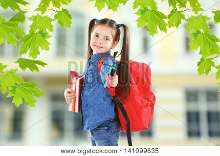 Little girl with red back pack holding books and stationery on blurred school building background
