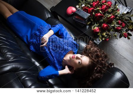 Young beautiful brunette girl lies on a leather couch and stares into the camera. Roses are on the table. Shooting from above.
