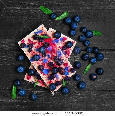 Chocolate bars White blackberries blueberries blueberry raspberry candied violet petals mint leaves candy heart on dark black background wooden surface empty place for text top view