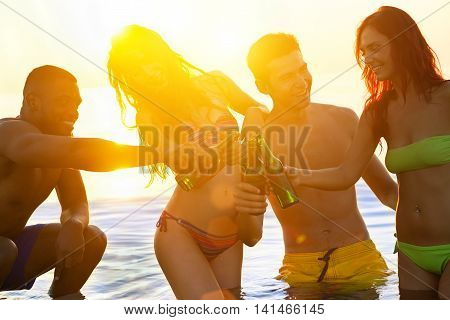 Cheerful friends sitting on their knees while having party on the beach at sunrise- Multi-ethnic young people toasting with beer bottles - Vacation and friendship concept - Sun original color tones