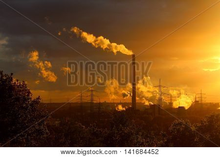 A power plant with a high chimney releasing white smoke and with an electrical line at the background at a mild-coloured sunset