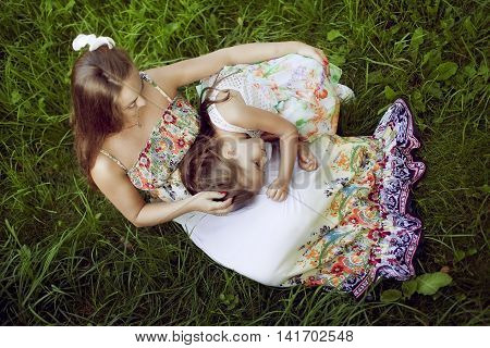 Mother and daughter in a beautiful floral sundresses in the summer sit on grass and hug. The girl clung to a pregnant woman and listens to her tummy. The view from the top