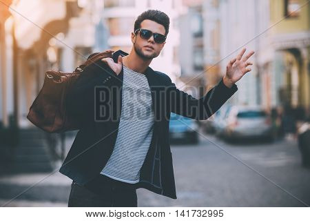 Taxi! Handsome young man in smart casual wear carrying bag on shoulder and gesturing while hitchhiking on the street
