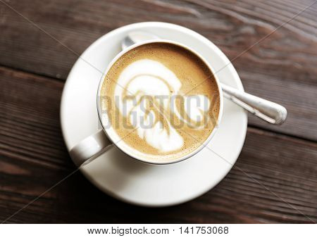 Cup of tasty latte is standing on the wooden textured table. There is nice picture is painted on the top of the latte. Latte is in the white big cup.