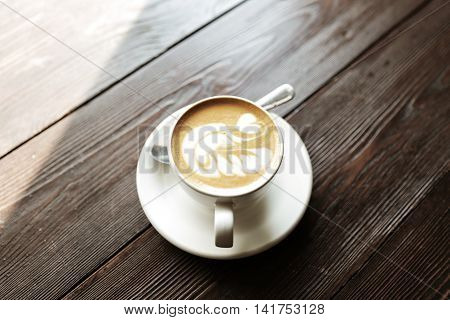 Cup of tasty latte is standing on the wooden textured table. There is nice picture is painted on the top of the latte. Latte is in the white big cup.