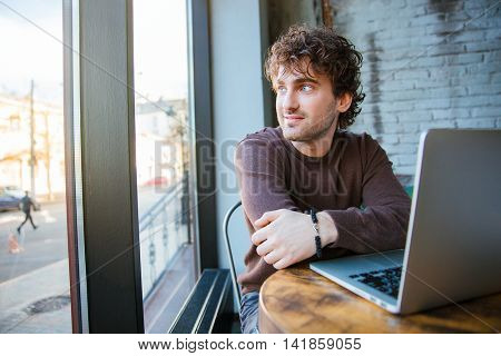 Curly handsome attractive content pensive happy young man sitting in cafe with laptop and enjoying the view