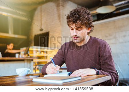 Concentrated thoughtful handsome attractive curly guy in brown sweetshirt planning his schedule sitting in cafe