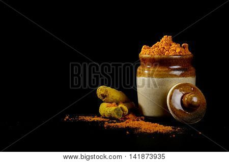 Turmeric powder in clay pot with roots or barks on black background