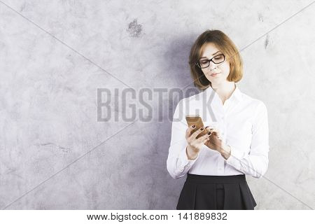 Attractive caucasian businesswoman in formal wear using smartphone against concrete wall with copy space