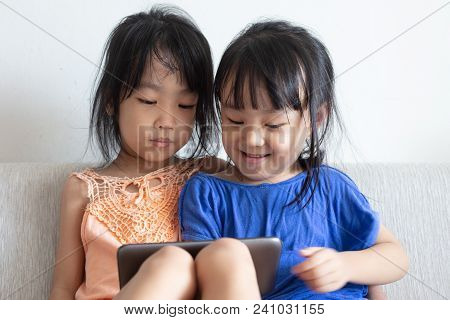 Asian Chinese Little Sisters Using Tablet On The Sofa In The Living Room