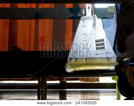 Urine Collection Bag Attached To Patient Bed With Orange Light From The Window In Hospital.